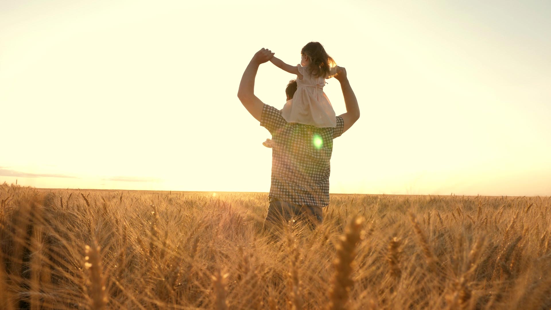 Farmer holding daughter on his shoulders in field during sunset